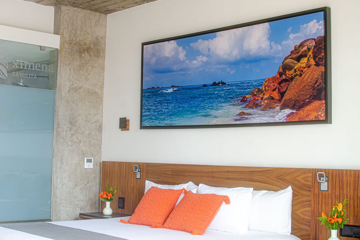 Habitación de hotel con una gran pintura con vista al océano, cabecera de madera y dos jarrones con flores.