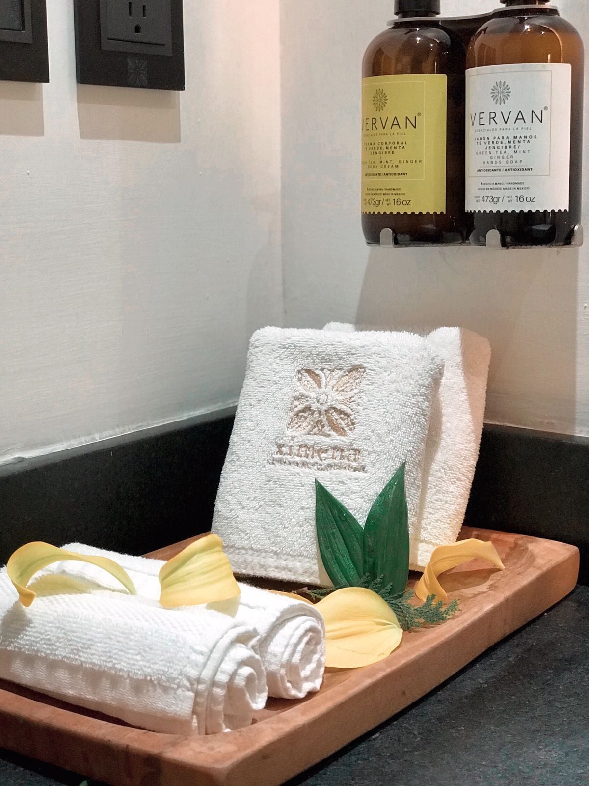 Ein Badezimmer mit einer Holztablett mit gerollten Handtüchern, einem weißen Handtuch mit floralem Muster und einem grünen Blatt. Über einer Ablage befinden sich zwei Flaschen Vervan Seife.