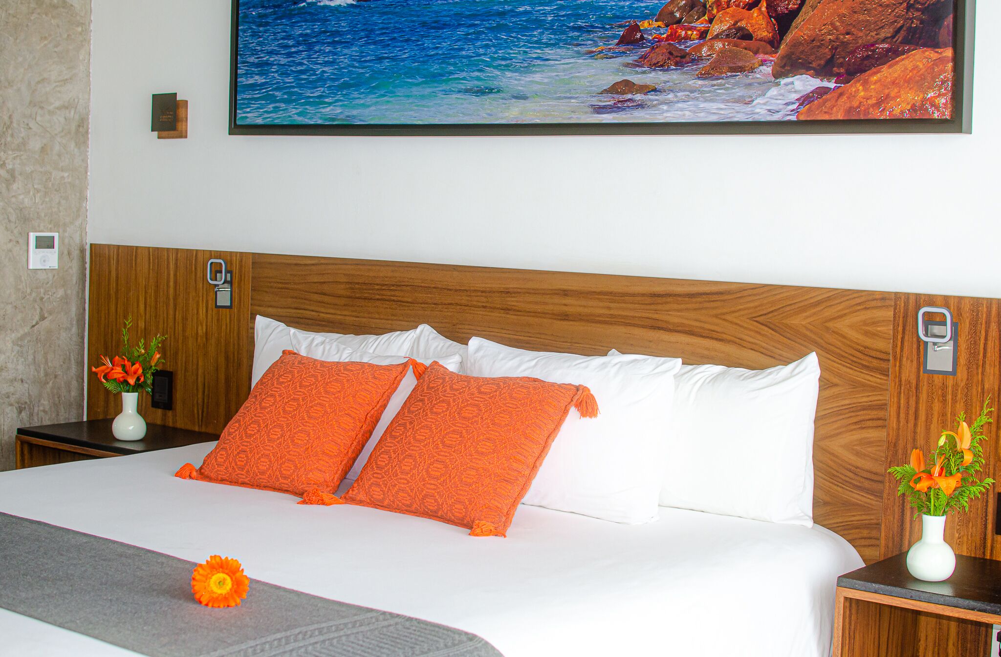 Habitación de hotel con cabecera de madera, ropa de cama blanca y almohadas naranjas. En la pared, un cuadro con una escena de playa y, en la mesita de noche, un jarrón con flores.