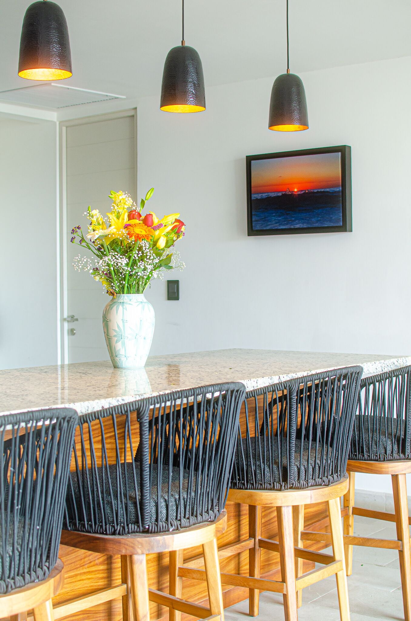 A dining area with a long wooden table, black woven chairs, and pendant lights.