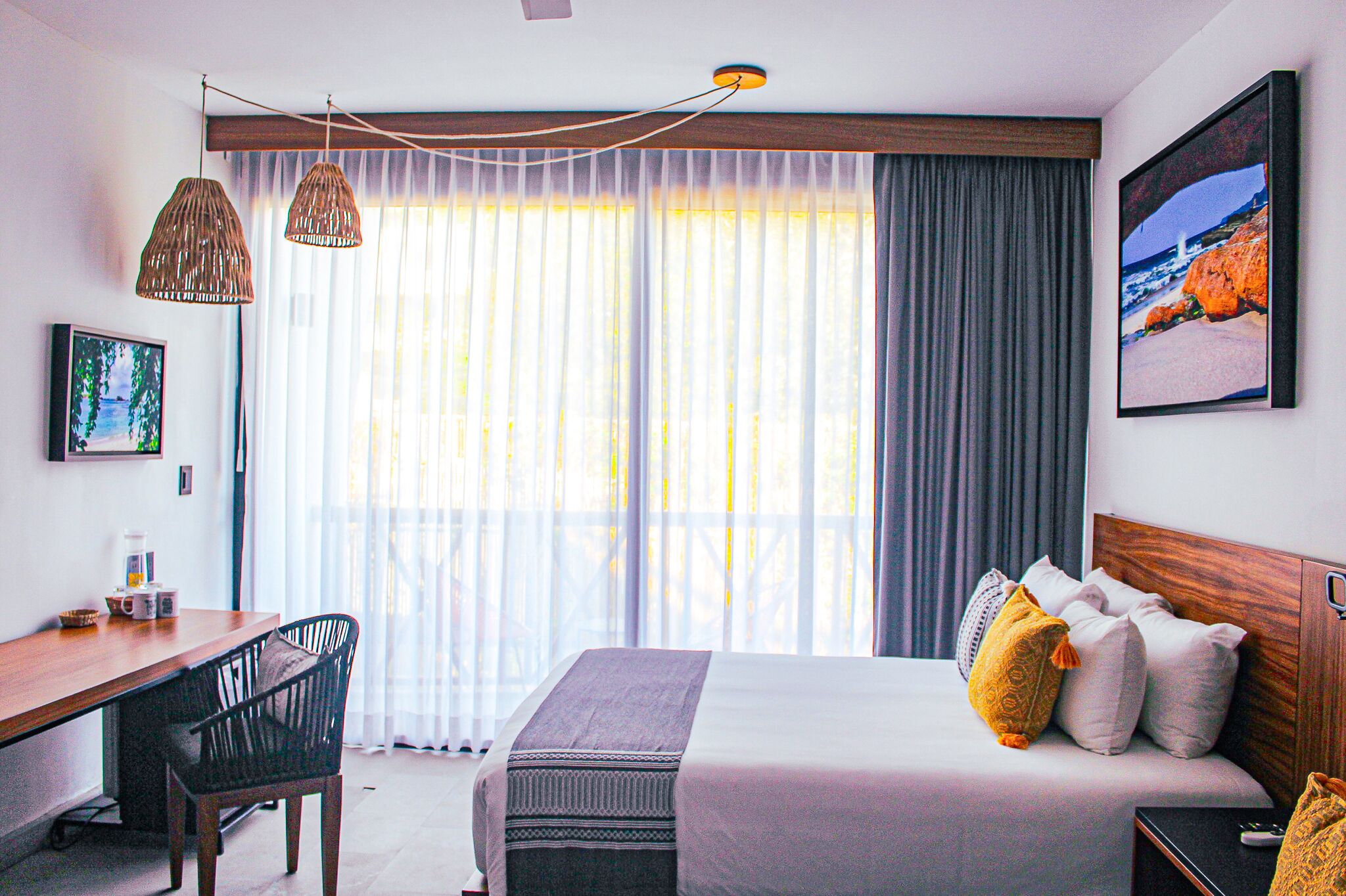 A hotel room with a bed, a desk, and a chair. There is a window with curtains, a picture on the wall, and a painting above the bed.