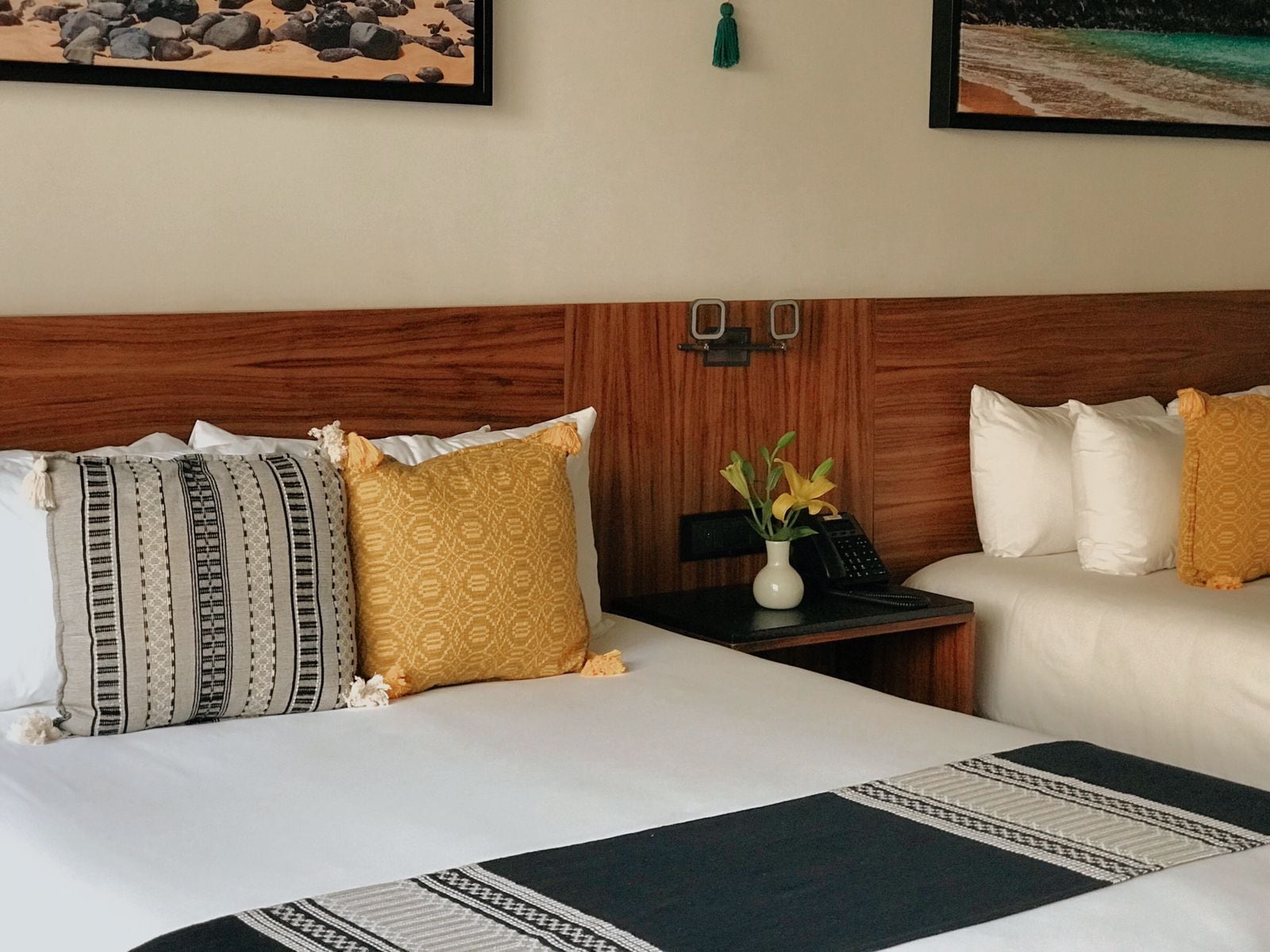 A hotel room with a wooden headboard, two beds with white and yellow bedding, a nightstand with a phone and vase, and two framed pictures on the wall.