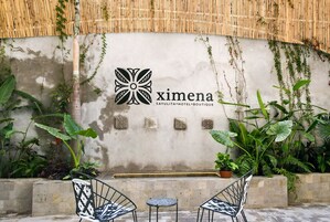 Un jardín con una pared de bambú, un espacio de estar con dos sillas y una mesita, y un logotipo de 'ximena' montado en la pared.