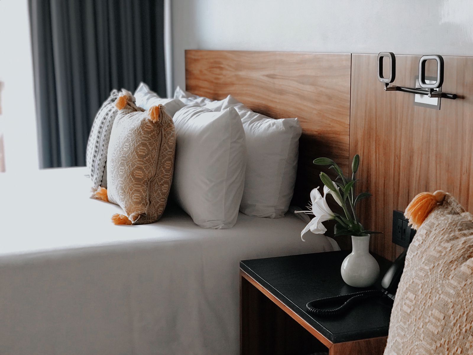 Una cama bien hecha con almohadas blancas y estampadas, una mesita de noche con un florero y un cabecero de madera.