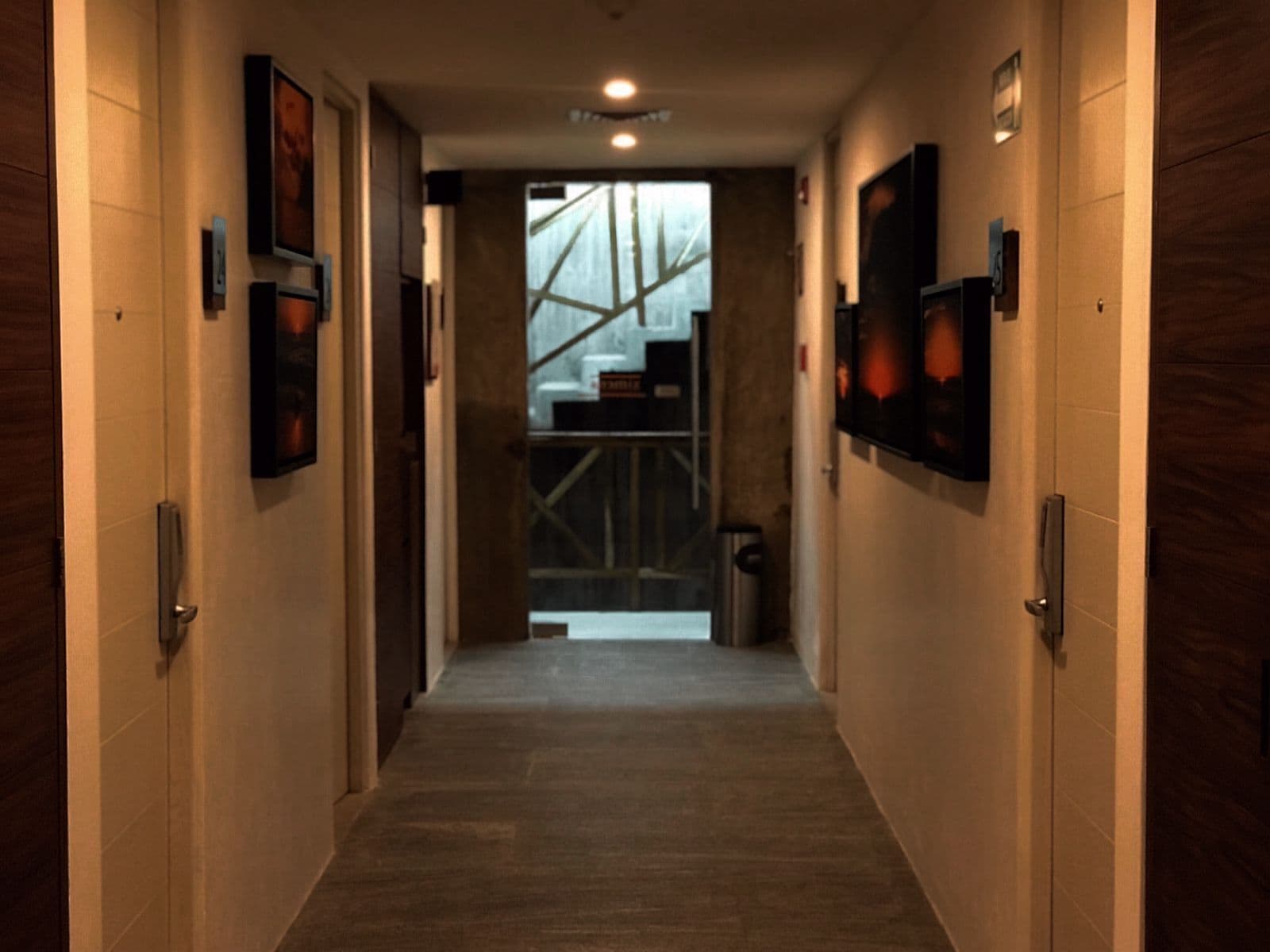 Een gang met ingelijste foto’s aan de wanden, een houten vloer en een zicht op een kamer met een raam en een metalen constructie.
