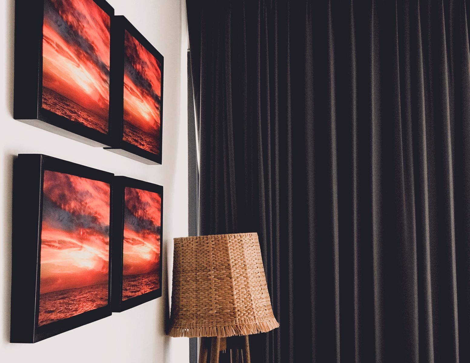 Una habitación con tres cuadros enmarcados de un atardecer, una cortina oscura y una lámpara de pie tejida.