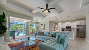 A spacious living room with a light blue sectional sofa, a glass coffee table, and a colorful rug. There is a kitchen area with white cabinets and stainless steel appliances.