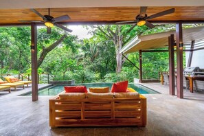 A wooden sofa with cushions in a covered outdoor area with a pool and natural surroundings.