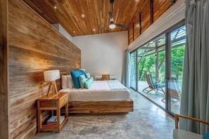 A modern bedroom with a wooden ceiling, a large bed, a nightstand, and a view of the outdoors through large windows.