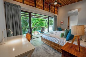 A bedroom with a large bed, a desk, and a chair. There is a balcony with a view of trees.