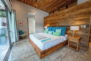 A modern bedroom with a wooden headboard, a large bed with patterned bedding, a bedside table with a lamp, and a view of a balcony.