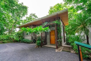 Una casa moderna con puerta de madera, rodeada de abundante vegetación y un camino de grava.