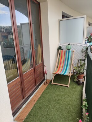Un balcon avec un revêtement de gazon synthétique, un hamac, une plante en pot et une porte en bois.