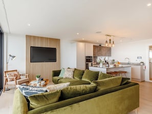 Un salon moderne avec un canapé vert, un téléviseur à écran plat fixé sur un panneau en bois et un coin cuisine doté d’un îlot central.
