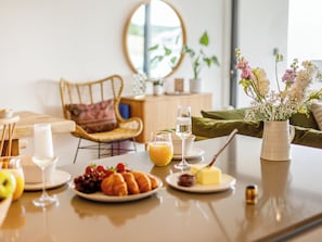 Frühstücksszene mit Croissants, Obst und Getränken auf einem Tisch, ein Korbstuhl und im Hintergrund eine Vase mit Blumen.