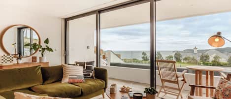 Un salón moderno con un sofá verde, una mesa redonda de centro y vista al mar a través de grandes ventanales.
