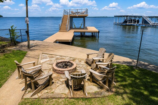 Una terraza de madera con sillas Adirondack, un pozo de fuego y un muelle que se adentra en un lago.