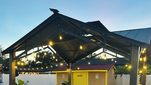 Uma área de estar coberta ao ar livre, com cadeiras e mesas, um edifício amarelo ao fundo e luzes pendentes.