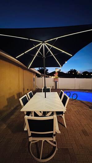 An outdoor patio with a table and chairs, a pool, and a decorative umbrella.