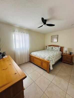 A bedroom with a bed, a nightstand, a ceiling fan, a window with curtains, and a wall-mounted picture.