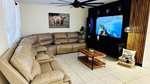 Uma sala de estar com sofá seccional, televisão de tela plana, ventilador de teto e um objeto decorativo na parede.