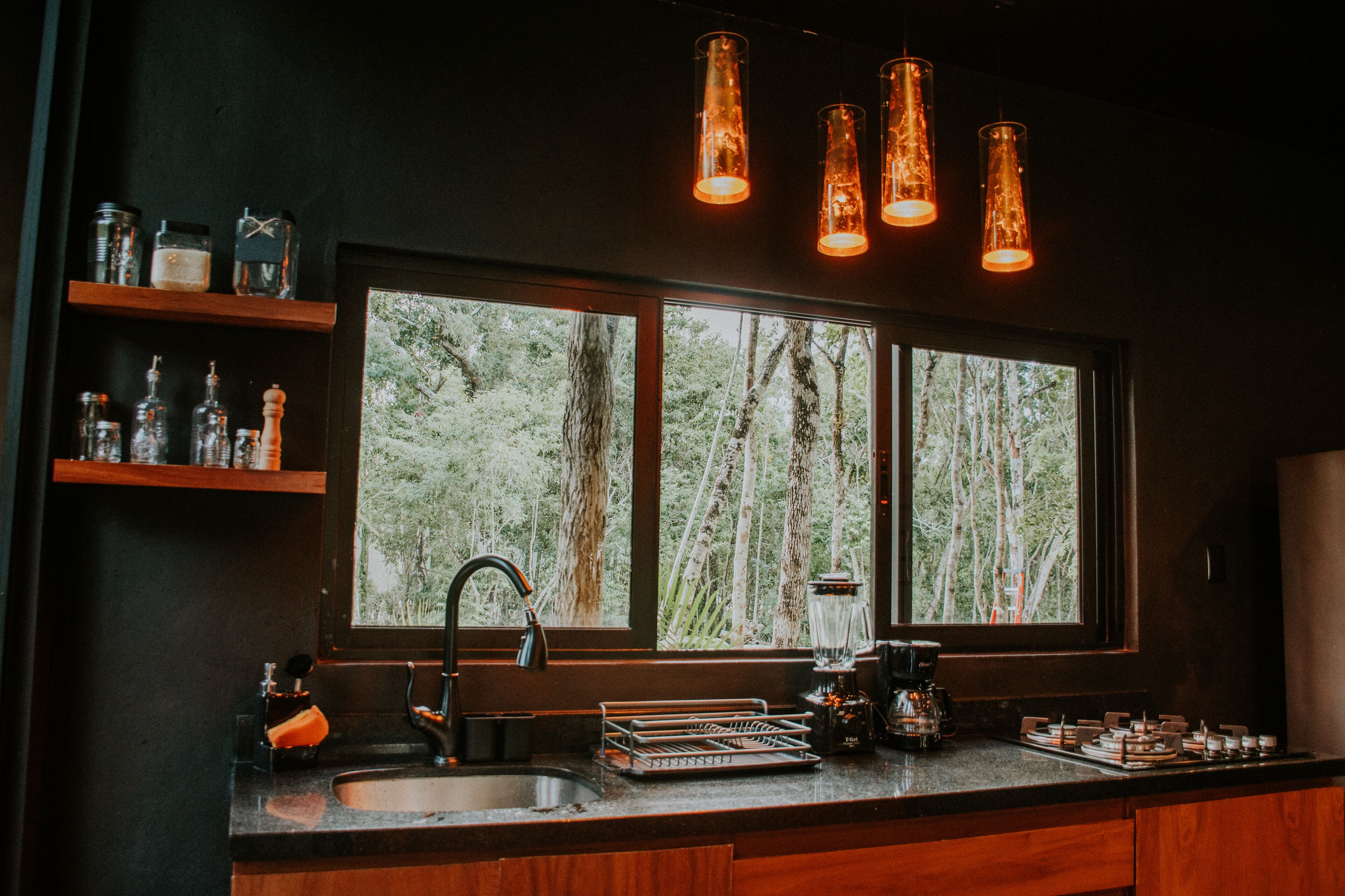 A kitchen with a large window, wooden shelves, and hanging pendant lights.
