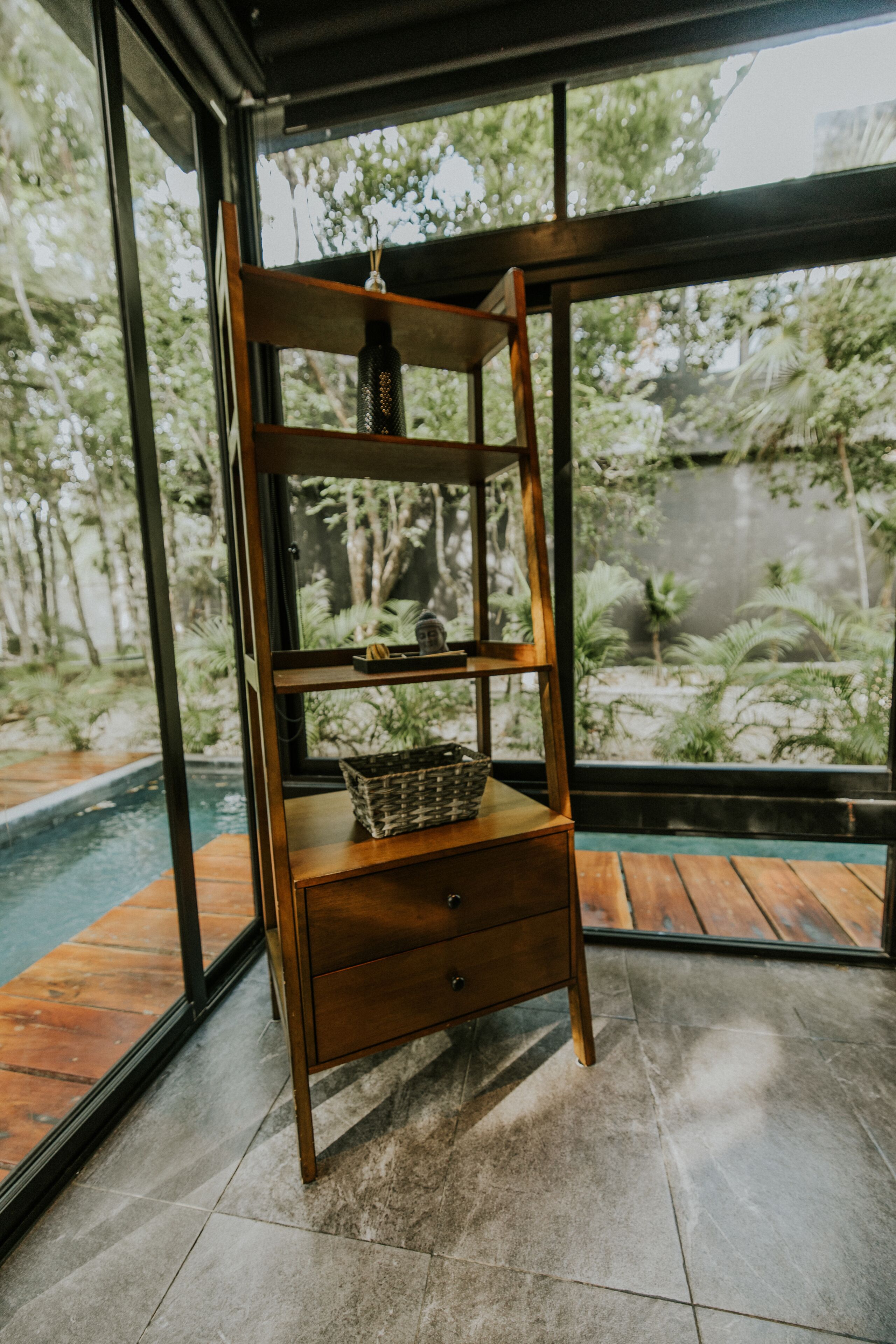 A wooden shelf with a basket and a small vase, placed in front of a large window overlooking a pool and greenery.