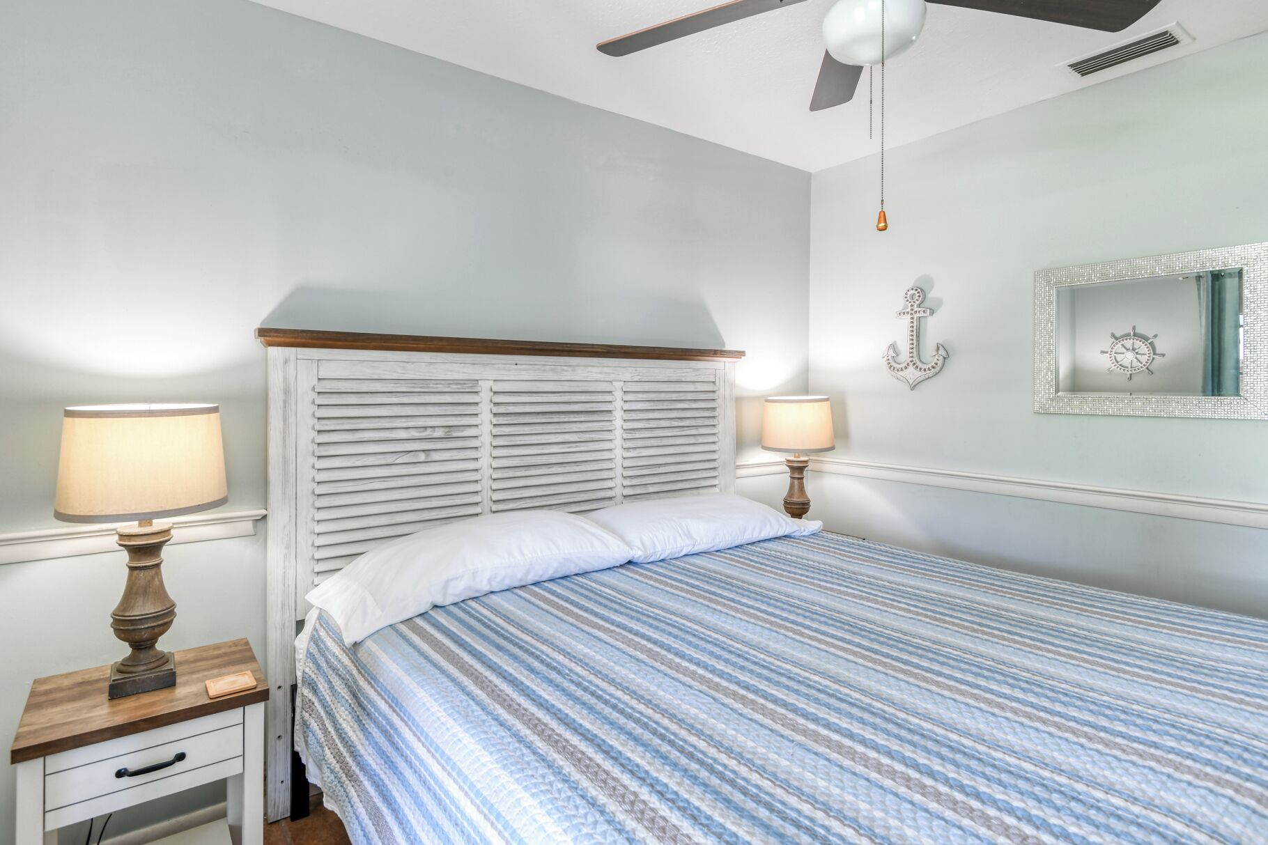 Un dormitorio con una cama, dos mesitas de noche con lámparas, un ventilador de techo y un ancla decorativa en la pared.