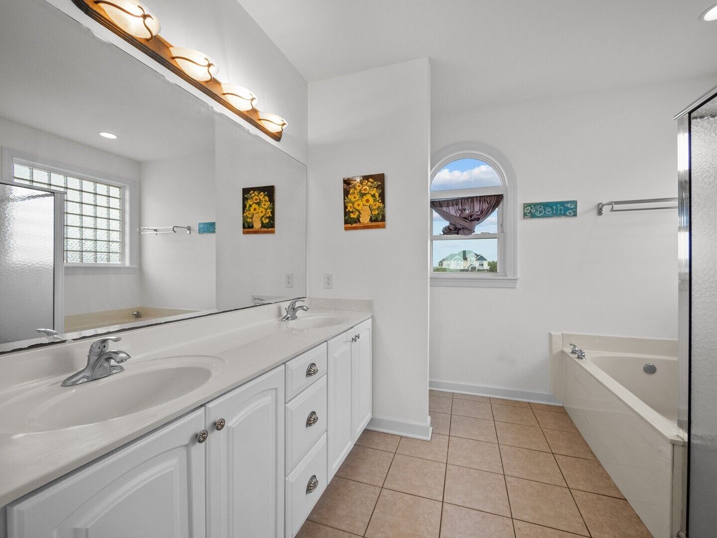 A modern bathroom with a double sink vanity, a large mirror, and a bathtub. There are two framed paintings on the wall and a window with a view of a house.