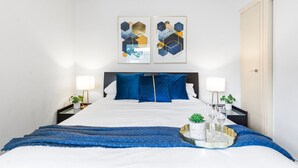 Une chambre d’hôtel bien aménagée, avec un grand lit, deux tables de chevet, une lampe de chevet, un plateau avec des verres et une plante, ainsi que deux tableaux abstraits accrochés au mur.
