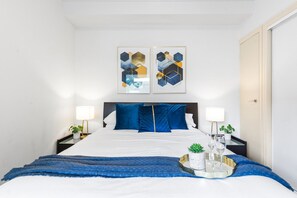A neatly arranged hotel room with a large bed, two bedside tables, a bedside lamp, a tray with glasses and a plant, and two abstract paintings on the wall. - The Cornerstone on Inkerman (St Kilda)