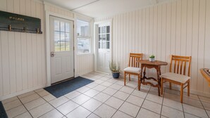 Una habitación con puerta de madera, una mesita con sillas, una planta en maceta y un felpudo azul.