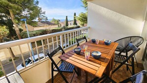 Une table en bois, entourée de quatre chaises, se trouve sur un balcon doté d’une balustrade blanche, d’où l’on a vue sur un quartier résidentiel avec ses maisons et ses arbres.