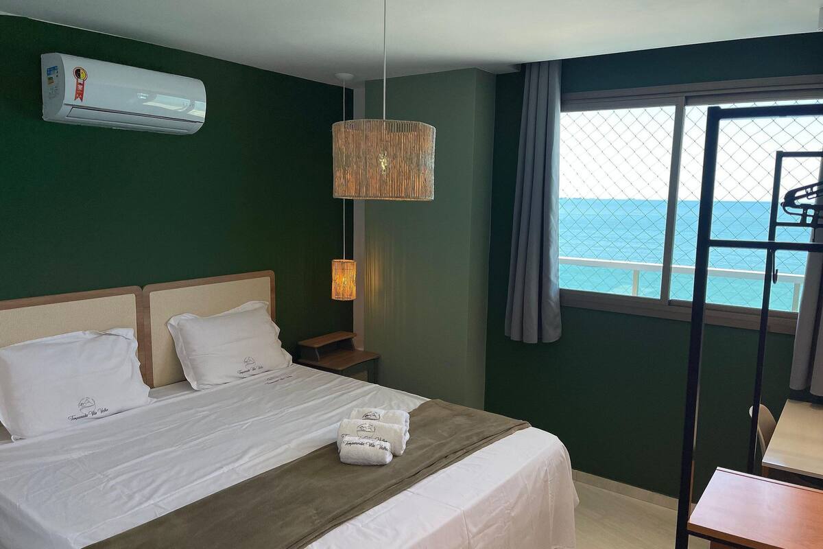 A hotel room with a bed, a nightstand, a hanging lamp, an air conditioner, and a window with a view of the sea.