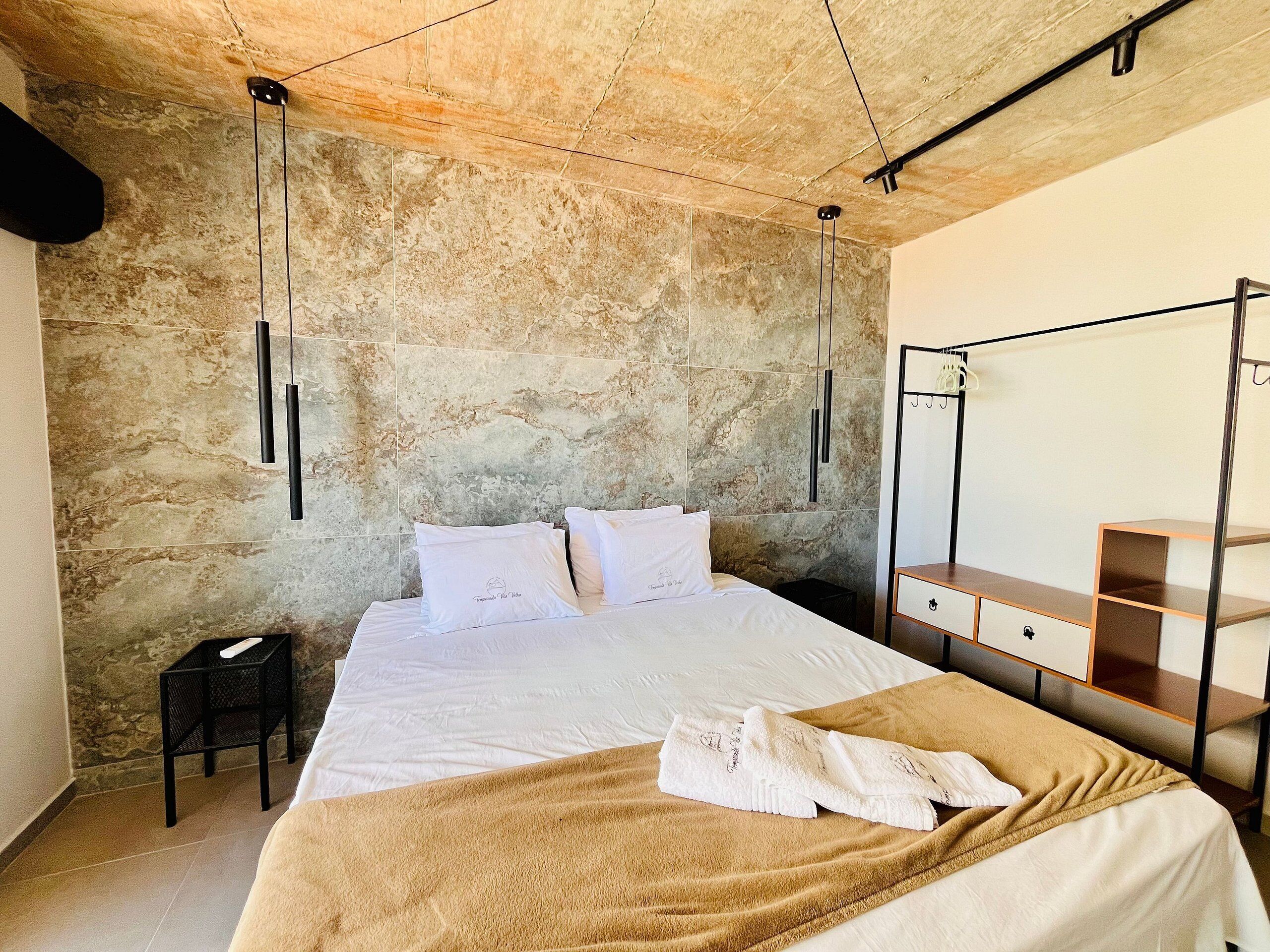 A bedroom with a large bed, a wooden nightstand, a mirror, and a stone-textured wall.