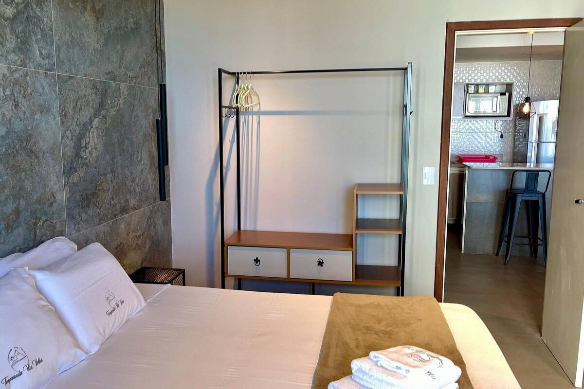 A bedroom with a bed, a wooden nightstand, a wardrobe, and a kitchenette in the background.