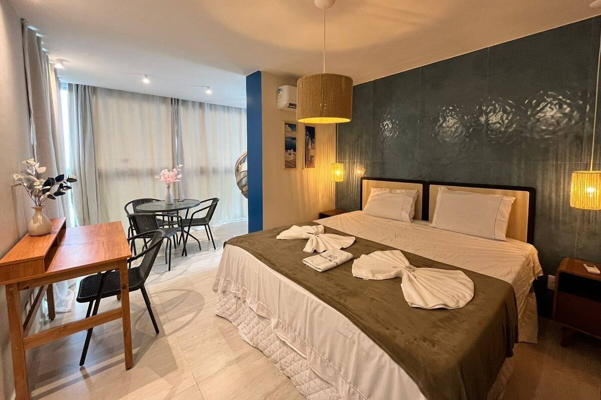 A hotel room with a bed, bedside tables, a desk with chairs, a dining table, and a large window with curtains.