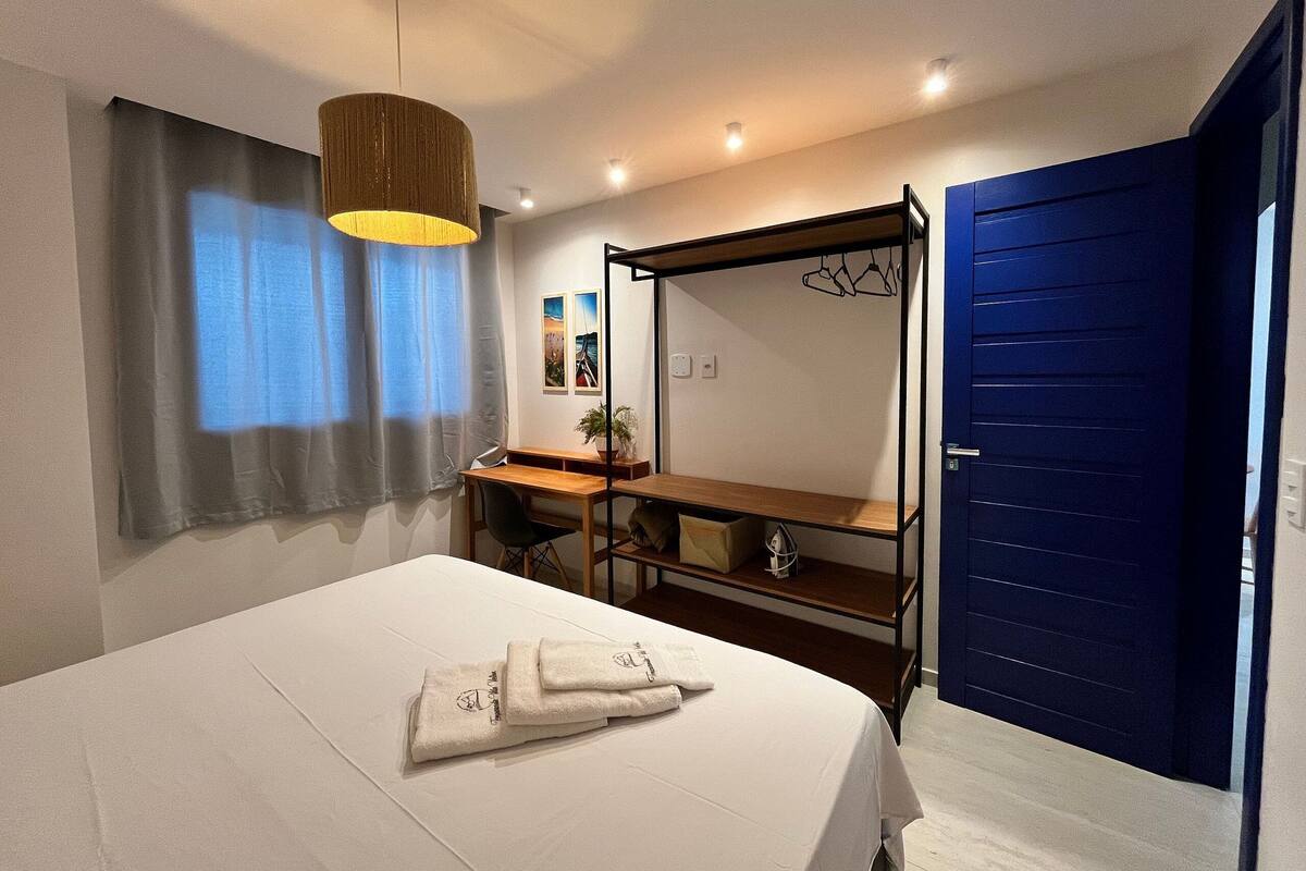 A bedroom with a bed, a desk, a chair, a hanging light fixture, a blue door, and a window with curtains.
