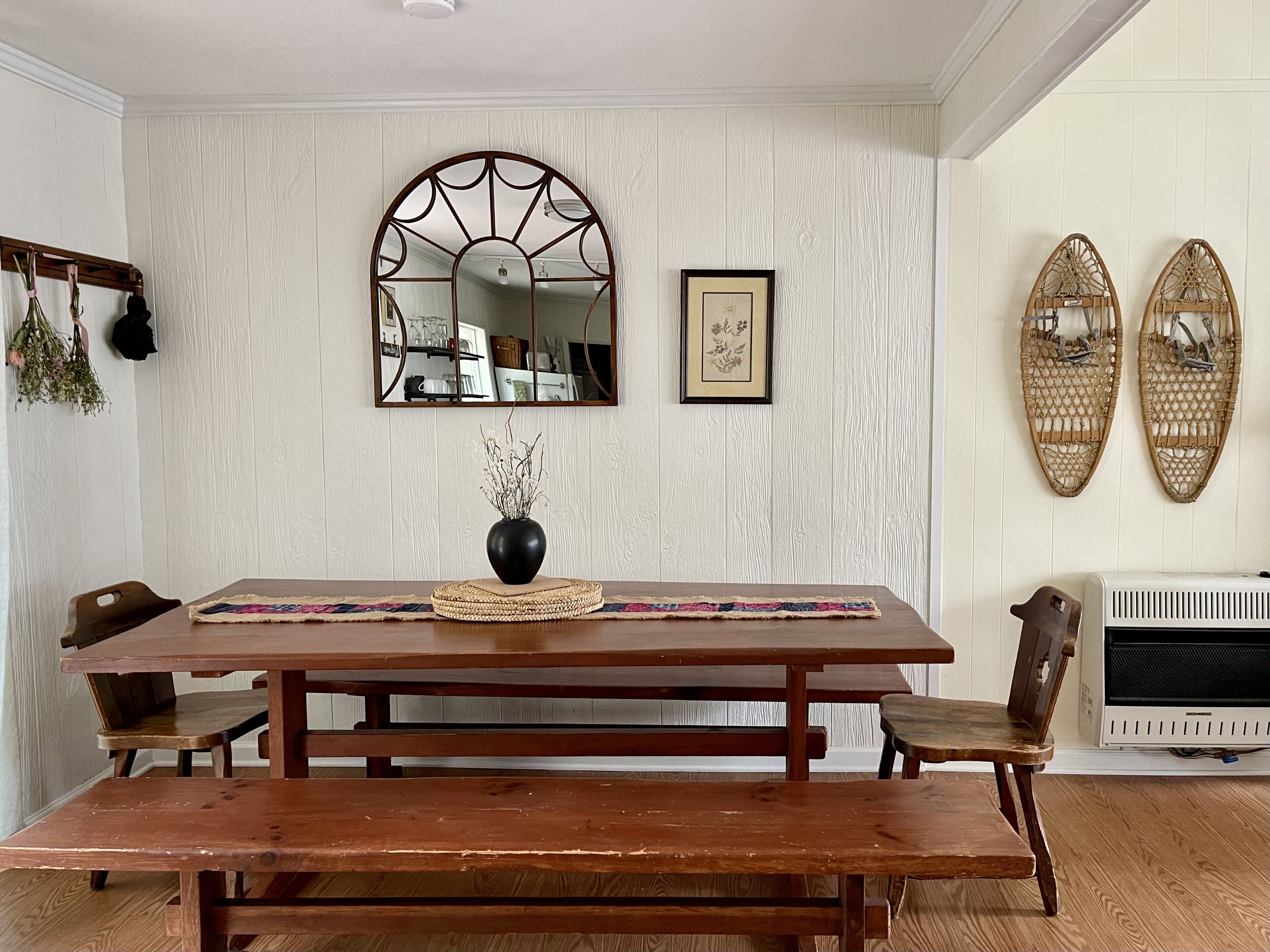 A dining area with a wooden table, benches, a vase with dried flowers, a mirror, and wall-mounted baskets.