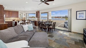 A spacious living area with a gray sectional sofa, a dining table with chairs, and a view of a body of water through large sliding glass doors. - 3526 -Bayside Walk Unit  2 (San Diego)