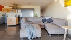 A modern living room with a grey sectional sofa, a small table, and a kitchen area in the background. - Luxurious beachfront, panoramic views (West Lakes Shore)