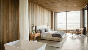A modern bedroom with a large bathtub, a bed with a view of the beach, and a wooden wall. - Luxury Designer Beachfront Home 3 Bed, 3.5 Bath in Arch Cape, Oregon. (Arch Cape)