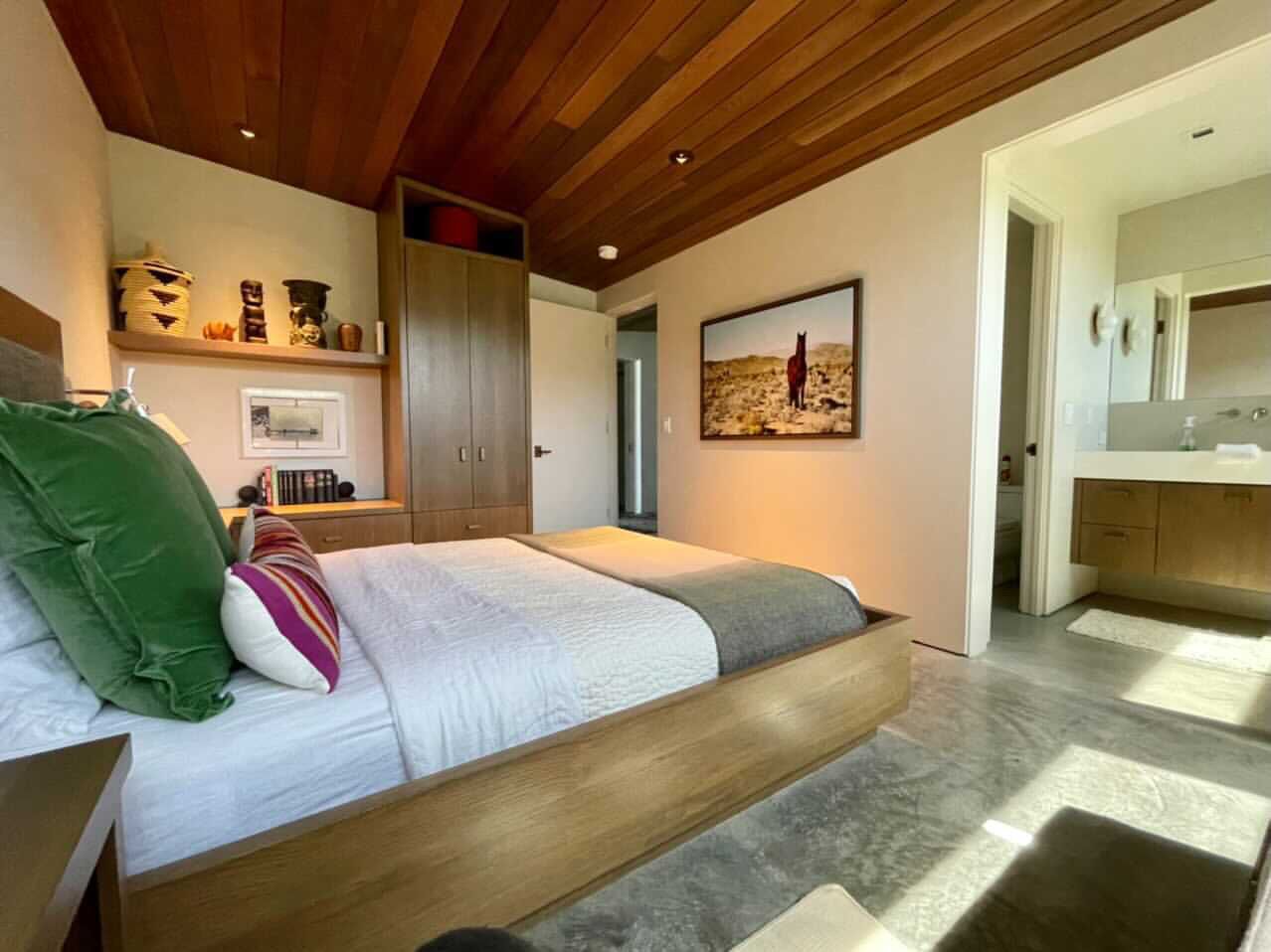A modern bedroom with a wooden ceiling, a bed with a green and striped pillow, a wooden nightstand, and a view of a kitchen and bathroom through an open door.