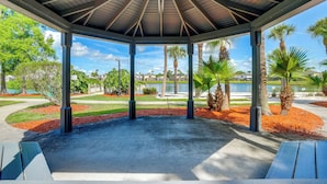 A gazebo with a concrete floor and a wooden roof, surrounded by palm trees and a water body.