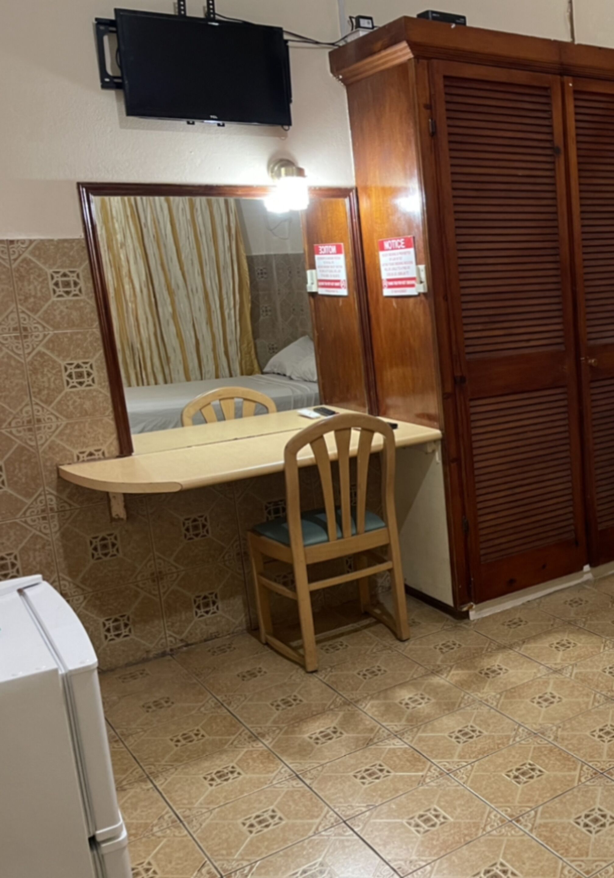 Una habitación pequeña con un armario de madera, una cama con cortina a rayas, una mesa de madera con una silla y un televisor montado en la pared.