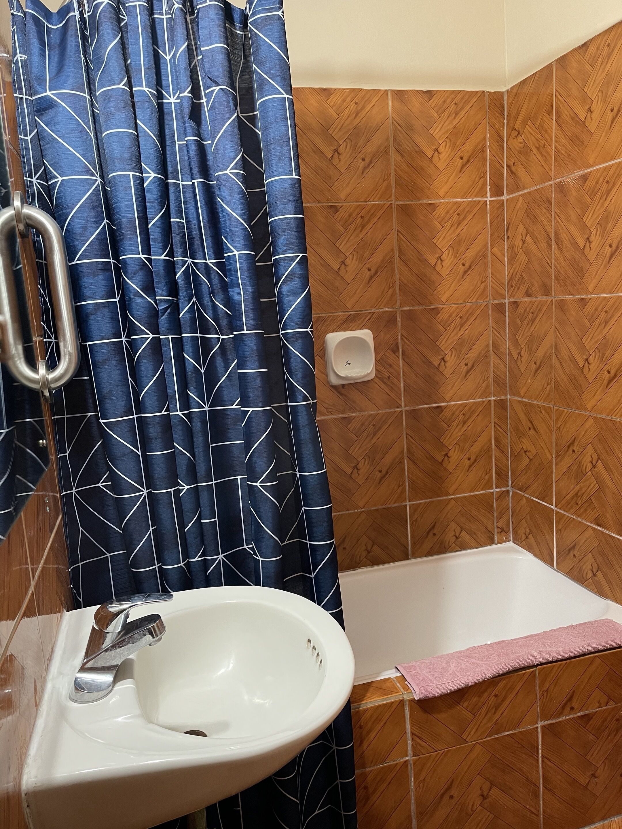 Un baño con un lavamanos blanco, una ducha con un patrón de espiga en madera y una cortina azul con diseño geométrico.