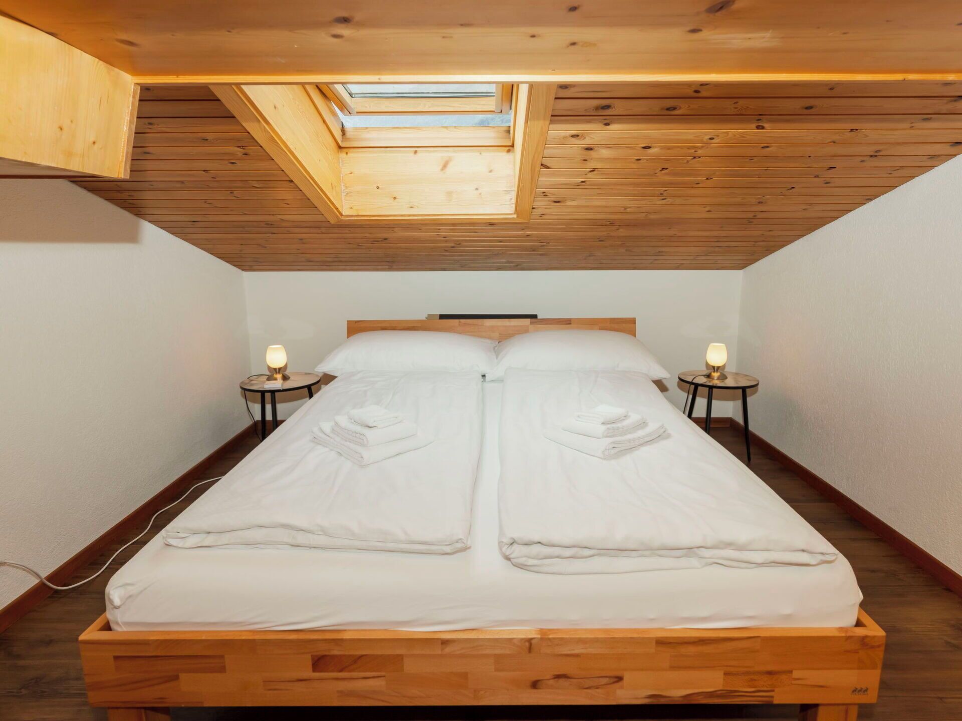 Un dormitorio con techo de madera, tragaluces, una cama con almohadas y lámparas de noche.