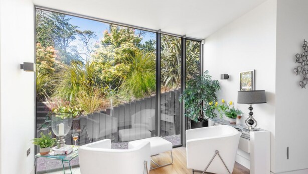 Un salon moderne doté d’une grande porte-fenêtre coulissante donnant sur un jardin, de meubles blancs et d’une petite table d’appoint avec une lampe.