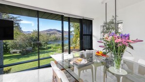 Un espace repas moderne avec une table en verre, des chaises blanches et une vue sur un lac et des montagnes.