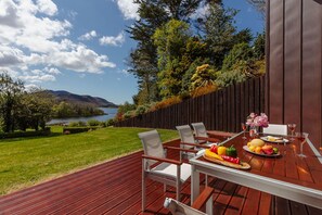 An outdoor dining area with a wooden deck, white chairs, and a table set with fruits and drinks.